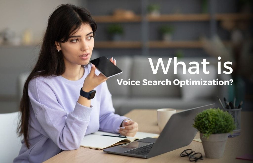 A young woman in a lavender sweater sits at a desk with her laptop, notebook, and coffee. She holds her phone near her mouth, using voice search. The background has shelves and plants. The text on the image says, "What is Voice Search Optimization?"